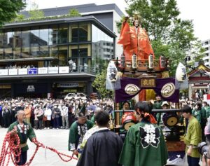 Crowds in happi coats carry an ornate gold mikoshi past office towers during the Kanda Matsuri shrine entry procession in 2023