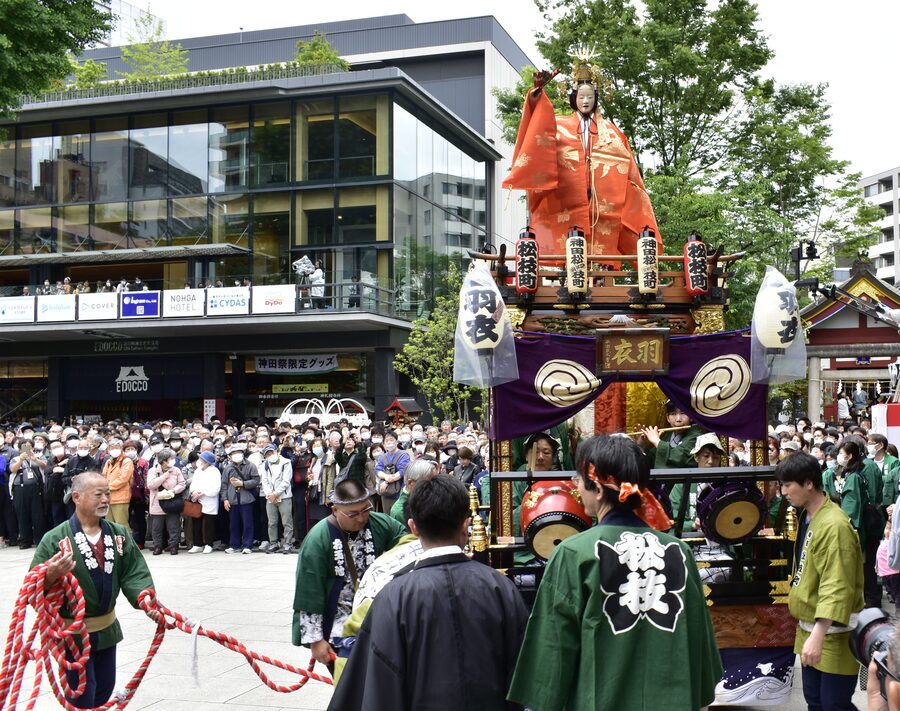Crowds in happi coats carry an ornate gold mikoshi past office towers during the Kanda Matsuri shrine entry procession in 2023.