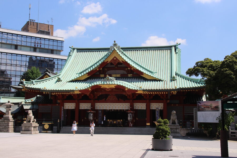 The vermilion grand hall of Kanda Myojin shrine with red lanterns visible during a calm day, and a glass office building visible in the background.
