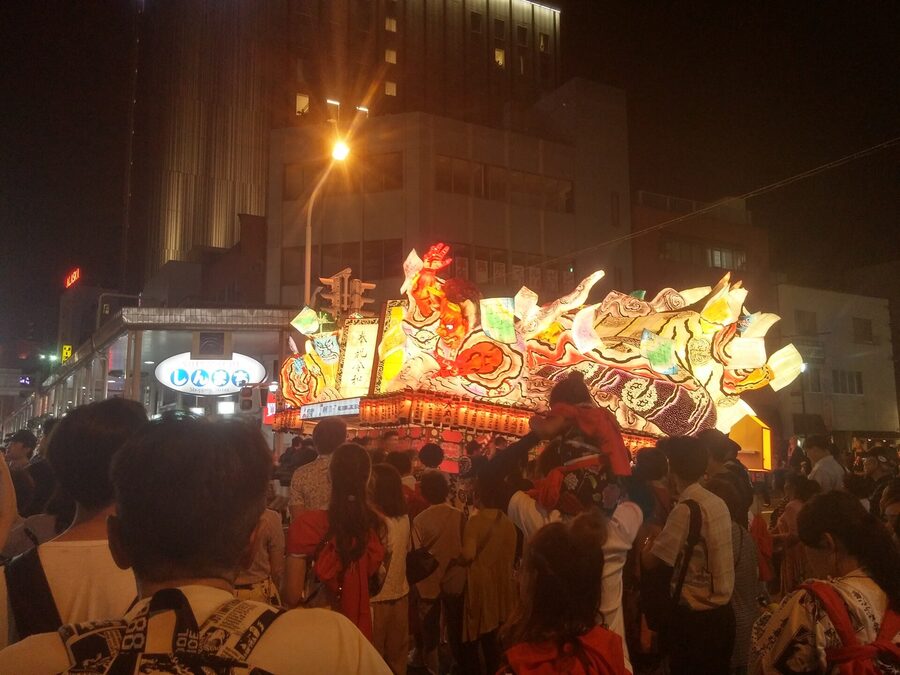 A samurai-themed Nebuta rolls down Shinmachi-dori with crowds packed against the barricades.