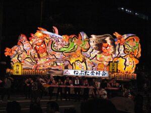 A massive painted Nebuta float lit from inside, depicting a warrior in motion against a dark sky over Aomori City.