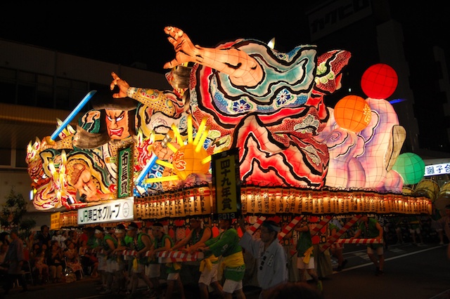The painted face of a warrior on a Nebuta float, lit from within by electric bulbs glowing through washi paper.