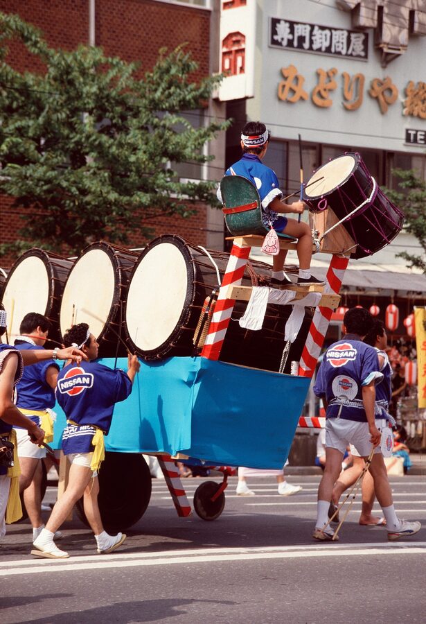 Haneto in straw hats and yukata leap through the parade route, bells jangling at hip and ankle.