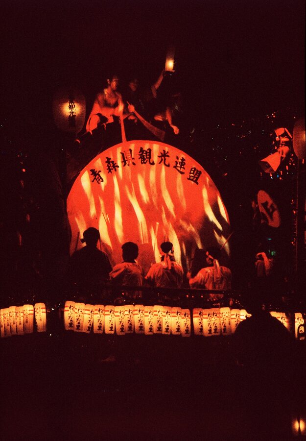 Taiko drummers ride a wheeled platform alongside a Nebuta, supplying the heartbeat for the haneto chant.