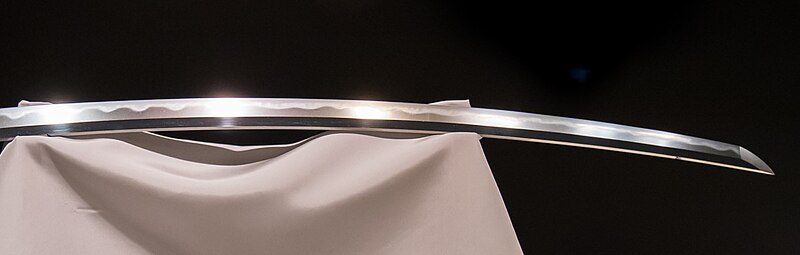 Close-up of a Japanese sword blade showing the hamon temper line a bright wavy band along the cutting edge contrasting with the smoky upper portion of the blade