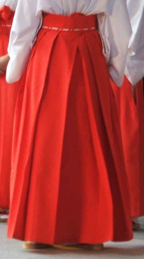 Miko shrine maiden from behind wearing traditional hi-no-hakama bright red hakama over white kosode at a Shinto shrine