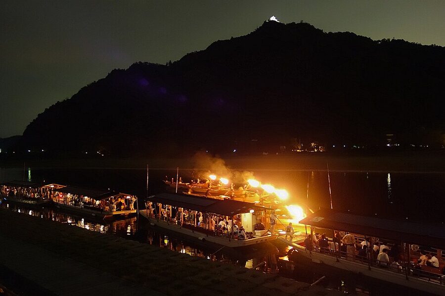 Ukai fishing master in traditional dark clothing working cormorants from the bow of a wooden boat, burning kagaribi torch illuminating the river, Nagaragawa Gifu